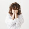 Studio portrait of tired or bothered woman hiding face in her curly hair and holding hands on it, expressing exhaustion or just being childish, standing over gray background. I am not seeing you